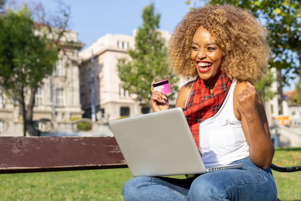 A smiling woman sitting on a bench outdoors holds a credit card and uses a laptop, looking excited and happy. Trees and buildings are visible in the background on a sunny day.