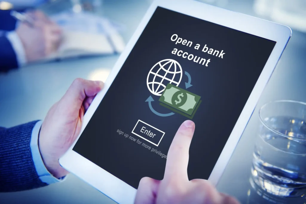 A person holds a tablet displaying a webpage to open a bank account, featuring a globe, dollar bill icon, and an Enter button. A glass of water and another person’s hands are visible in the background.