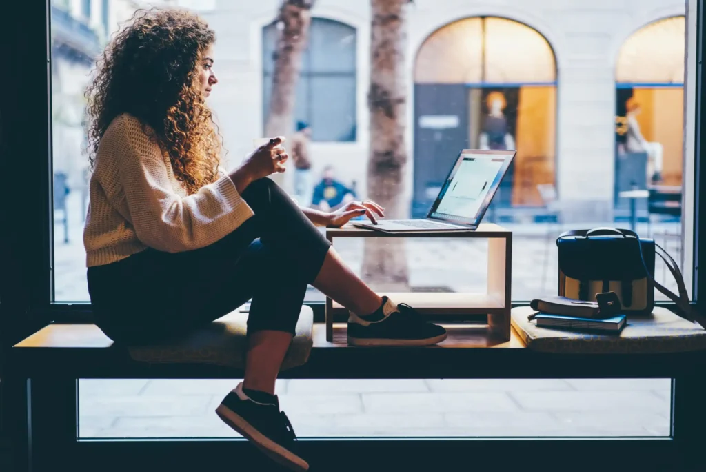 A woman with curly hair sits by a large window in a café, working on a laptop. She holds a cup and appears focused. A bag and notebooks are next to her, and city buildings are visible outside.