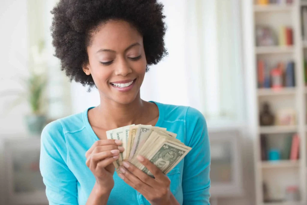 9 Best Rewards Checking Accounts of March 2026 A smiling woman in a blue top counts a stack of U.S. dollar bills, standing indoors with soft light and shelves blurred in the background.