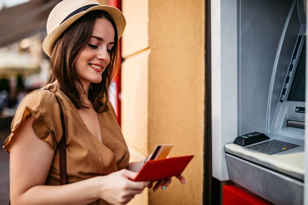Best Free Checking Accounts in March 2026 A young woman wearing a tan dress and straw hat smiles while holding a wallet and bank card, standing next to an outdoor ATM machine.