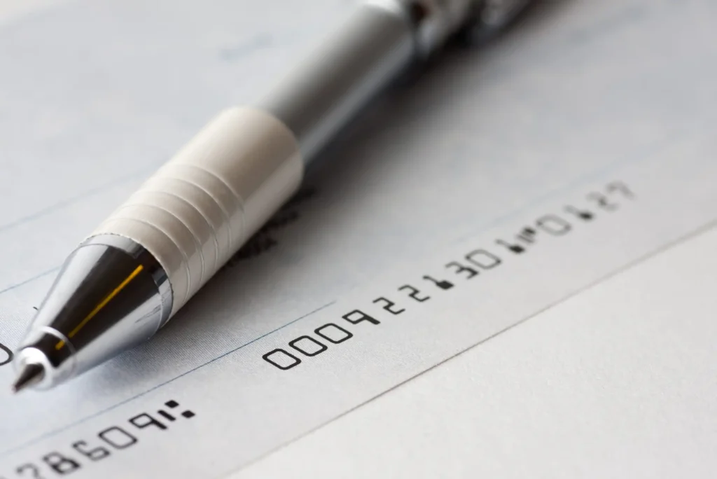 A close-up of a silver and white pen resting on a blank check, with visible numbers and a faint signature line.