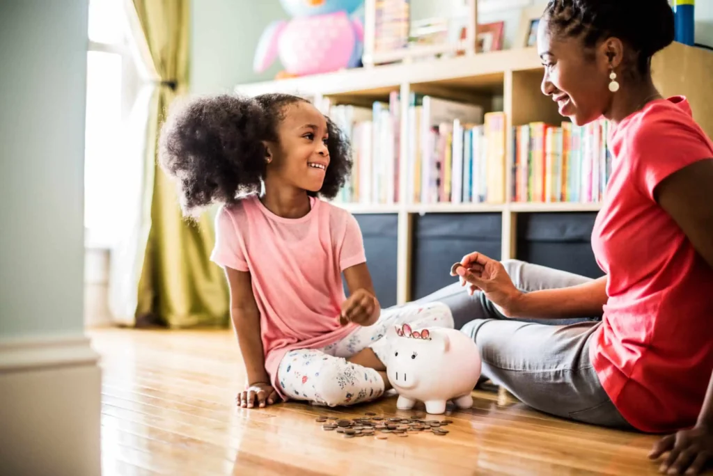 Best High-Yield Savings Accounts for March 2026 A smiling young girl and an adult sit on the floor, counting coins next to a piggy bank in a bright room with bookshelves and toys in the background.