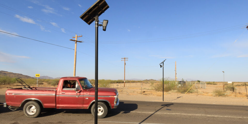 A red truck carrying a lawnmower passes two Flock Safety ALPRs attached to poles.
